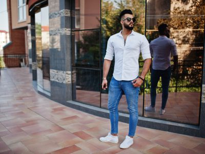 Stylish tall arabian man model in white shirt, jeans and sunglas Stylish tall arabian man model in white shirt, jeans and sunglasses posed at street of city. Beard attractive arab guy against modern building.