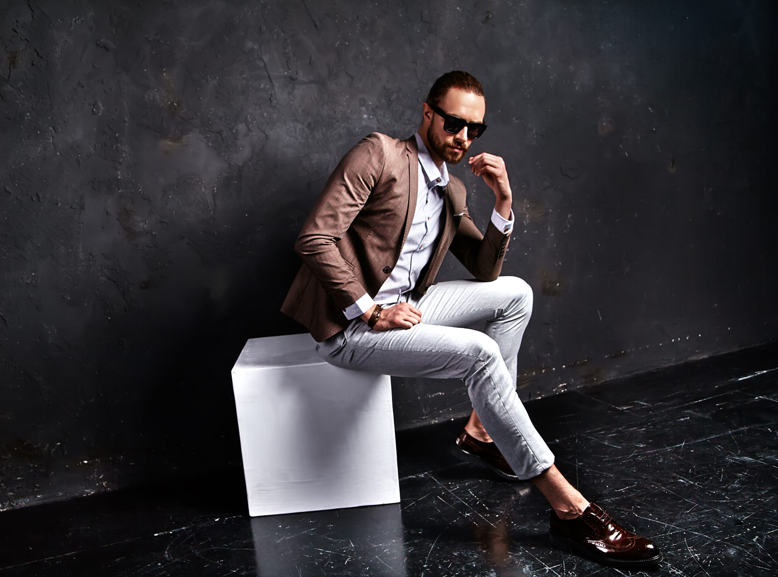portrait of handsome fashion stylish hipster businessman model dressed in elegant brown suit sitting near dark wall in studio in sunglasses
