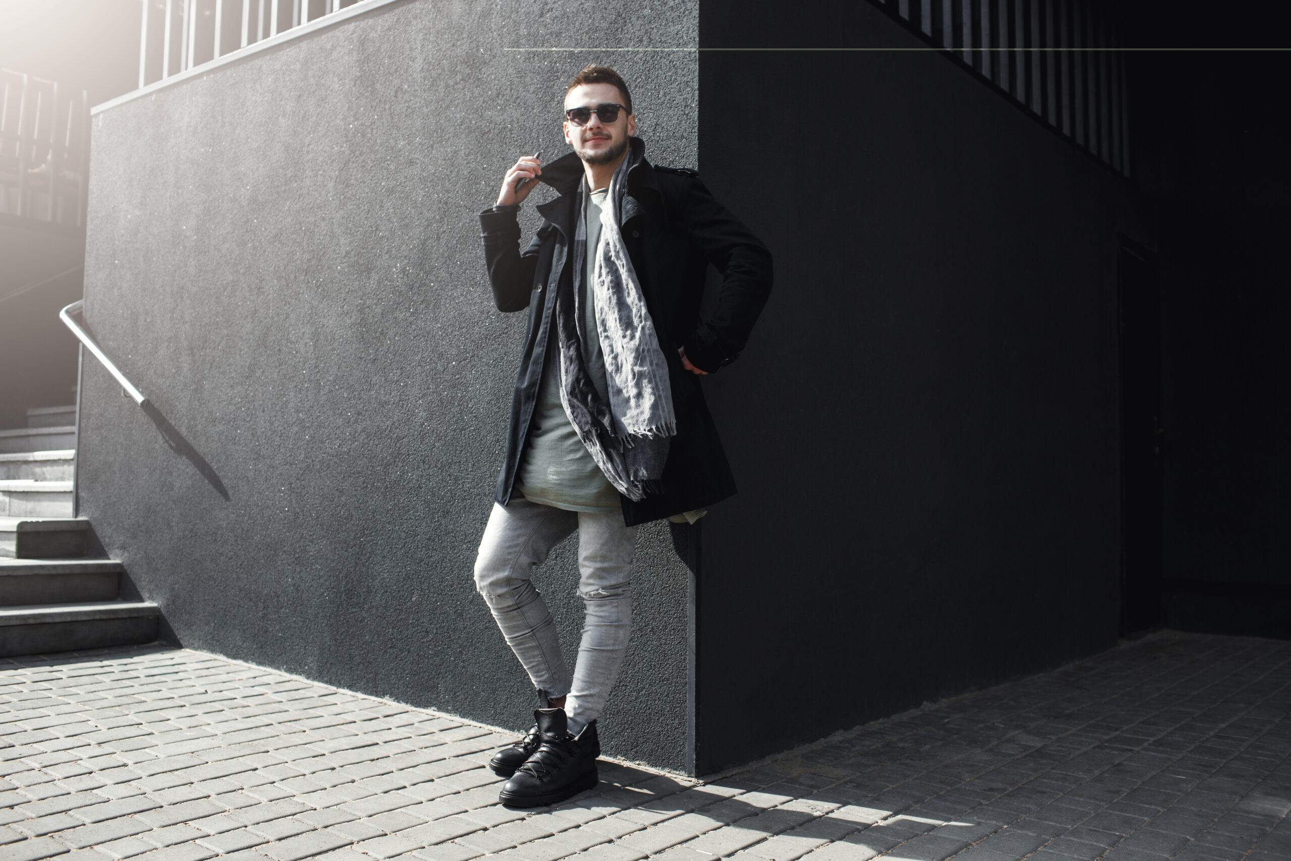 Nice guy in stylish clothing standing outside, leaning to wall, holding phone, looking away.