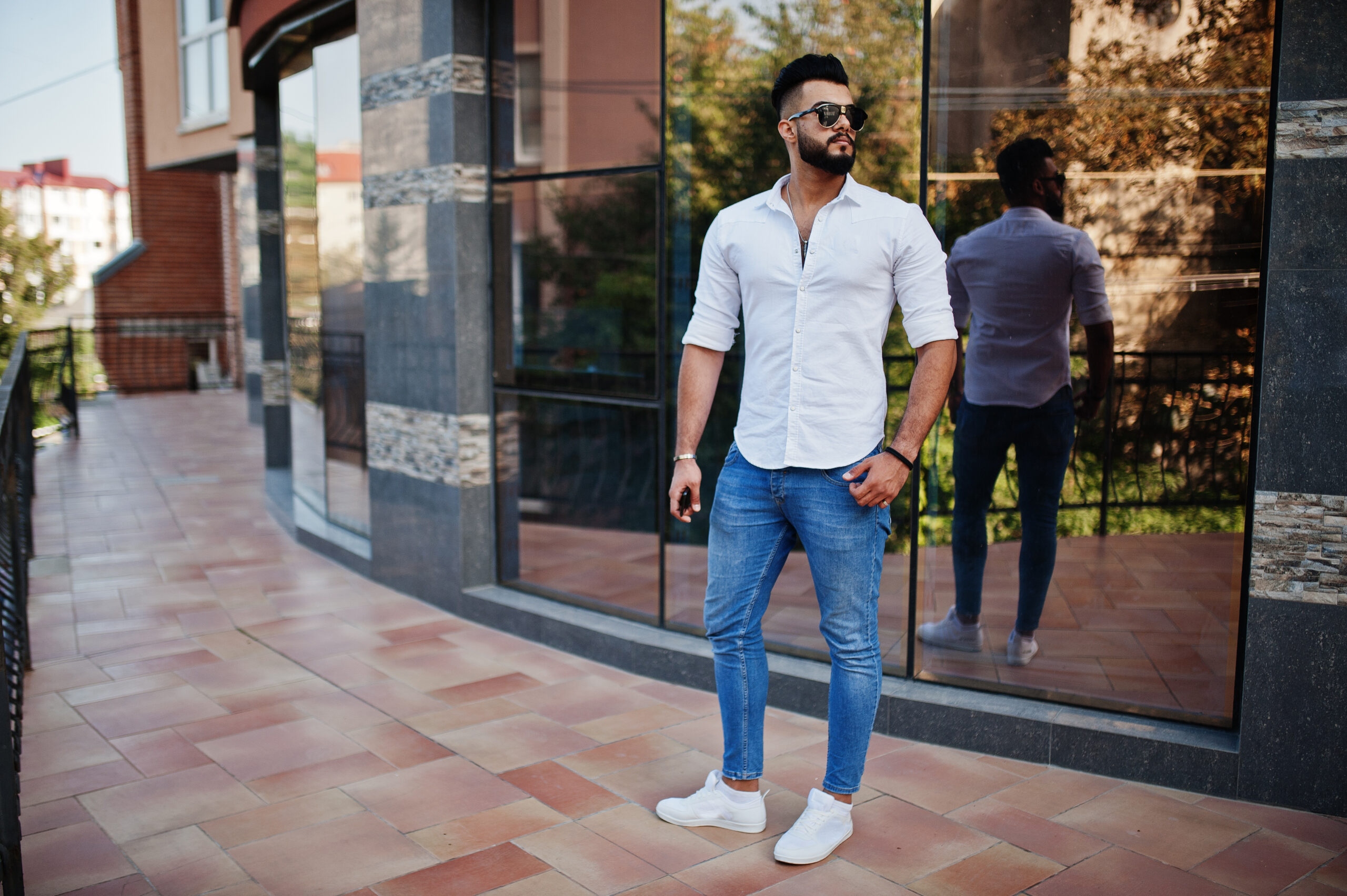 Stylish tall arabian man model in white shirt, jeans and sunglasses posed at street of city. Beard attractive arab guy against modern building.